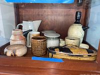 Full view of art pottery collection including sake set, pitcher, vases, and lidded box in a wooden cabinet.