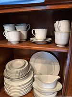 Full view of stacked dinner plates, luncheon plates, soup bowls, cups on shelves, showing embossed floral pattern on cream-colored Grace Concerto china.