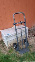 Photo showing metal dolly/hand truck in front of outdoor storage trunk outside against building.