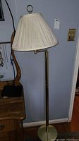 Photo of standing floor lamp with pleated cream shade by a wall and wooden furniture.