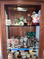 Wood cabinet shelf with an assortment of glassware including clear and green-tinted glasses, copper mugs, ceramic Asian figurines, ginger jars, and vases