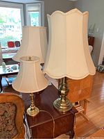 Three brass table lamps with different cream fabric shades shown on wooden furniture with living room background.