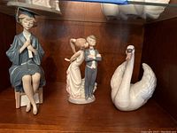 Three Lladro figurines on wooden shelf: seated graduate girl, kissing boy and girl, swan figurine.