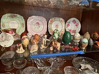 Shelf display showing Royal Albert tea cup and saucer with assorted Royal Stafford plates behind; various decorative eggs and small dog figurines in front; clear glass pieces on lower shelf