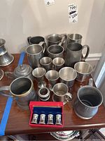 Photo of multiple pewter mugs, cups, a sugar bowl, and a red box containing four salt and pepper shakers arranged on a wooden surface.