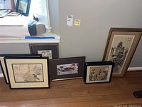 Nine framed Asian style artworks leaning on floor and windowsill showing variety of sizes and frame styles.