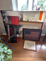 Photo showing various framed wall art leaning against a wall including a signed and numbered print by Pisani, an American flag print, and abstract paintings.