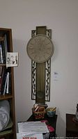 Full view of the tall, slender metal wall clock with round face and pendulum, mounted vertically on wall next to bookshelf.
