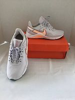 Front and side view of the light gray and peach Nike Legend women's shoes on white surface with orange Nike box underneath.