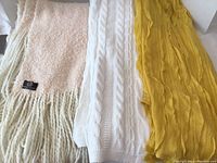 Photo showing the three throw blankets laid out side by side, featuring a yellow cable knit throw, a white cable knit throw, and a textured peach and white throw.