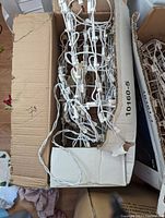 Box of white metal Christmas light frames with white string lights in a cardboard box, showing multiple connected bulbs and wiring.