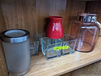 Shelf with four glass items including a frosted glass canister, a clear square compartment vase, a red rounded vase, and a large clear jar with metal lid and handle