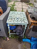 Outdoor metal table shown from a side angle, detailing the intricate patterned top and straight legs. Shows weathered light green paint and surrounding outdoor setting.