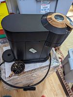 Side and top view of black Nespresso coffee machine showing circular copper control dial and removable drip tray.