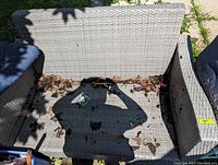 Wicker style outdoor love seat frame in light gray with no cushions. Seat has leaves and debris inside. Taken from above at an angle.
