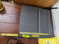 Paper chopper is shown closed with a grey base featuring measurement grids and rulers, a yellow handle for cutting, and a safety warning sticker.