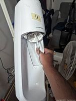 Photo showing front of white SodaStream carbonation machine with nozzle and carbonation tube visible, partial hand holding the unit, fan and toys in background.