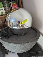 Photo showing a textured gray resin basin supporting a large smooth metallic reflective sphere, with attached electrical cord for lighting and pump.