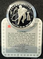 Front side of coin with hockey player and Stanley Cup design, displayed over certificate with details about the coin's significance, silver content, diameter, and weight.
