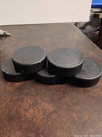 Five black rubber hockey pucks arranged side by side on a brown surface showing the tops and edges.