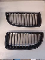 Pair of gloss black kidney grille inserts laid flat on white surface, front view showing vertical slats and shape.