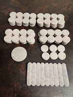 Large collection of round and rectangular beige felt pads with adhesive backing arranged in stacks by size on a dark surface.