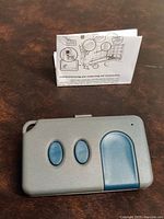 Gray programmable garage door remote control with three blue buttons displayed alongside an instruction manual for setup and compatibility.