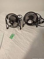 Photo showing two small individual black electric fans side by side on a white surface with power cords visible.