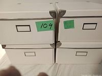 Four white rectangular reinforced boxes stacked in two rows showing metal corner protectors and label holders on the front sides.
