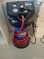 Front angle showing Campbell Hausfeld 2 gallon air compressor, blue tank, black top with gauges, red hose coiled on bottom.