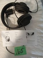 Photo showing black Fisher Bluetooth headphones with attached cable and instruction manual lying on a fabric surface with a green card marked 165.