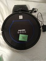 Top view of circular black Kalorik robot vacuum cleaner with blue trim, labeled buttons, charging station, and power adapter cord seen.