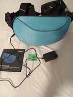 Photo showing the bluish-green massage unit with black straps, connected to a black power adapter, alongside the user manual.