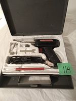 Open black plastic carrying case with white molded foam insert containing an electric soldering gun, power cord, two soldering tips, a small wrench, and a red handled tool.