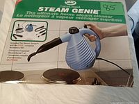 Photo showing the front of the product box with Steam Genie steam cleaner and accessory kit images, highlighting the blue device and black attachments.