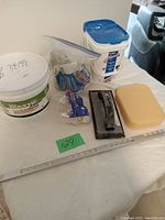 Full lot of tiling supplies including glue, grout, spacers, edge strip, grout float, sponge arranged on white cloth surface.