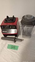 Red Vitamix blender motor base with dial controls, container with lid, and tamper placed next to it on white cloth.