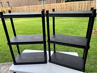Two black plastic shelves with three tiers each shown outdoors on a white table.