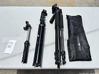 Photo showing two black camera tripods, a monopod, and a black carrying bag on a white table.