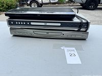 Three DVD players stacked on a table outside, top one slim with Blu-ray logo, middle a Magnavox DVD player, bottom an Emerson DVD player.