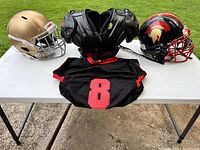 The lot displayed on a white table outside on concrete ground featuring two helmets, shoulder pads, and a jersey.