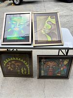 Four framed art pieces displayed on outdoor table featuring abstract faces in varying artistic styles and colors