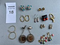 Top view of assorted earrings arranged on a grey table surface showing multiple pairs: gold hoops, floral dangling, colorful studs and drop earrings.
