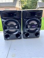Pair of two black Sony stereo speakers shown side by side on an outdoor surface with grass and buildings in the background.