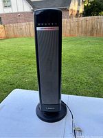 Full front view of the black Lasko ceramic tower space heater placed outdoors on a gray surface with a lawn in the background.