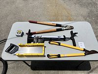 Overview of assorted hand and cutting tools on a white table, clearly showing handles, blades, and metal components.