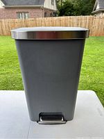 Front view of trash can showing rectangular shape, gray body, stainless steel lid, and foot pedal at the base.