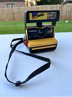 Front angled view of Polaroid Job Pro 2 camera showing label, lens, viewfinder, flash, and strap on white surface outdoors.