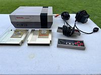 Overview of NES console, two game cartridges, one controller, power adapter, and AV cable on white surface outdoors.