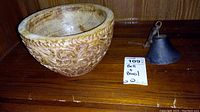 Photo showing the carved wooden bowl and the metal bell side by side on a wooden surface with the lot tag visible.